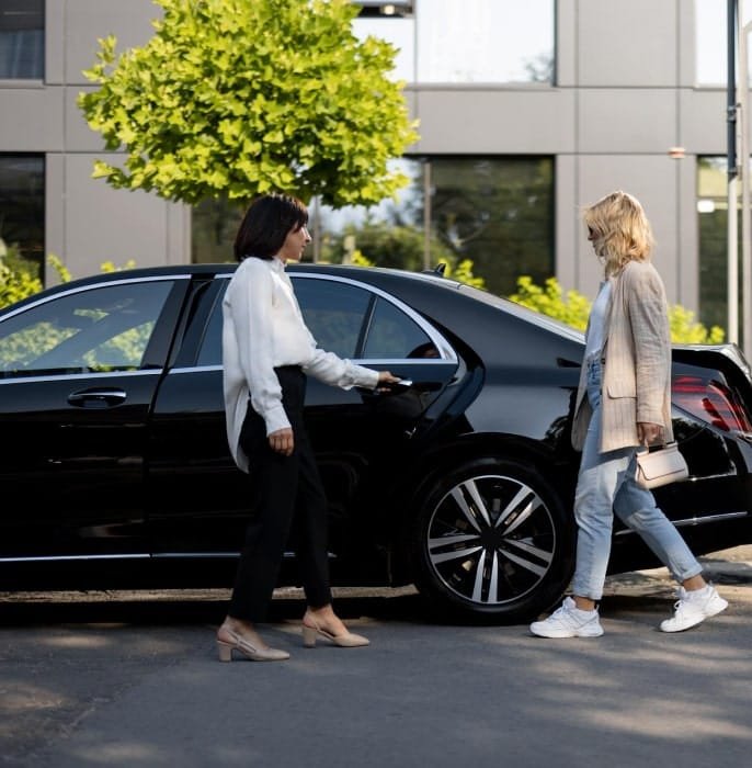 Two women standing beside a dark sedan outside modern buildings