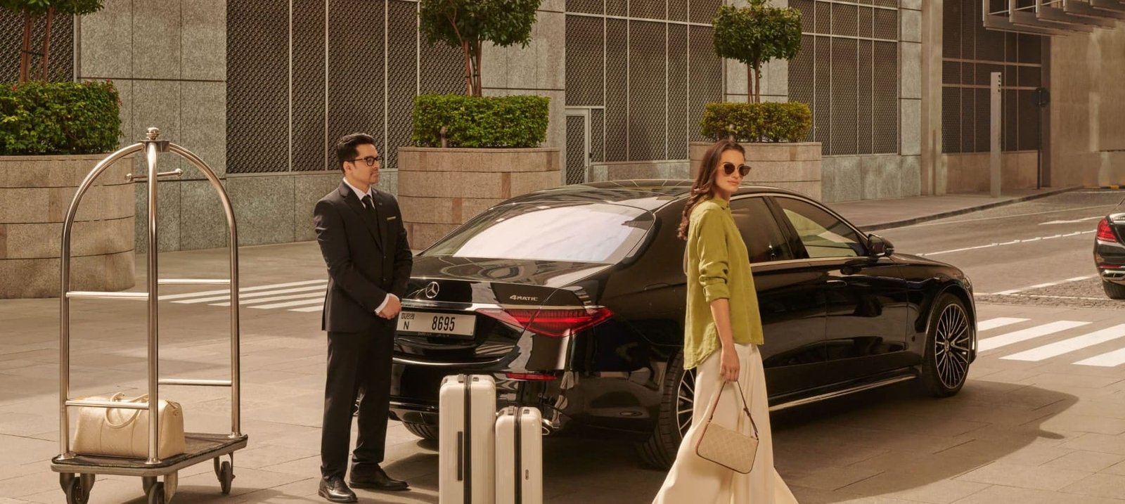 Travellers beside a black saloon and a hotel luggage trolley