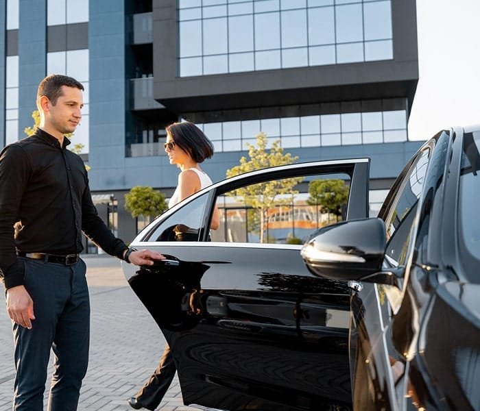 Man holding an open car door as a passenger approaches outside a glass-fronted building