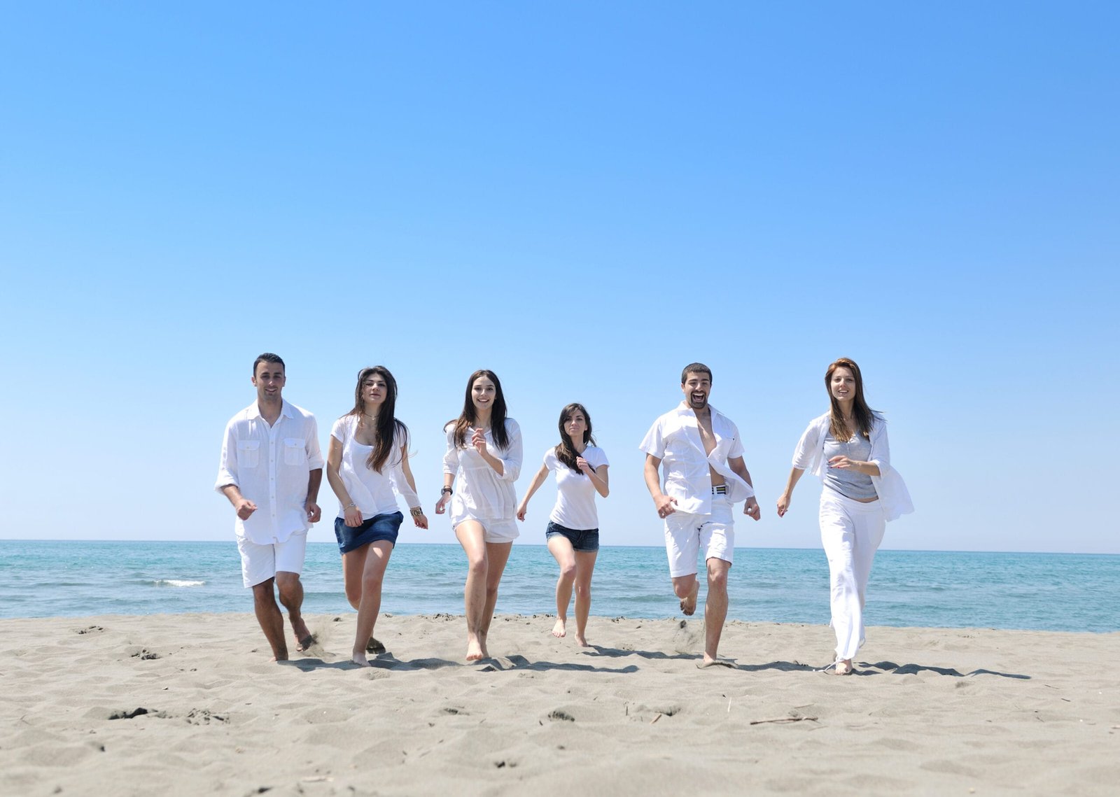 Six friends in light summer clothing running towards the camera on a sandy beach under a clear blue sky