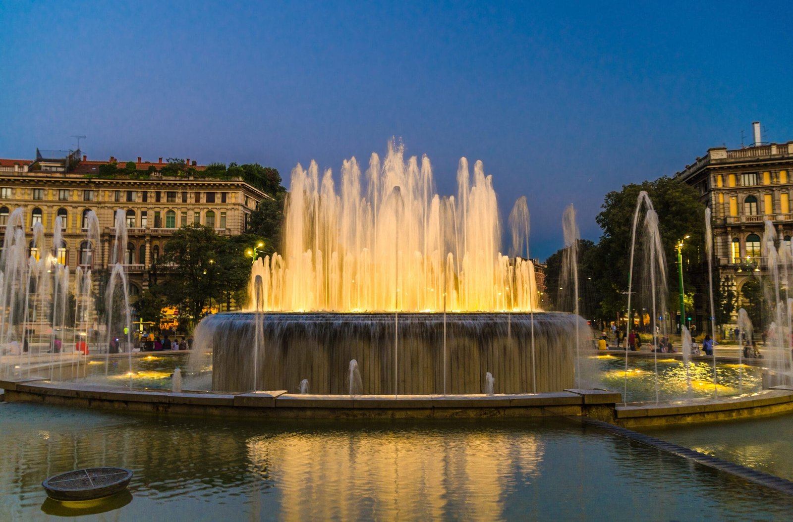 Illuminated fountain in Piazza Castello, Milan, with a blue evening sky and surrounding buildings