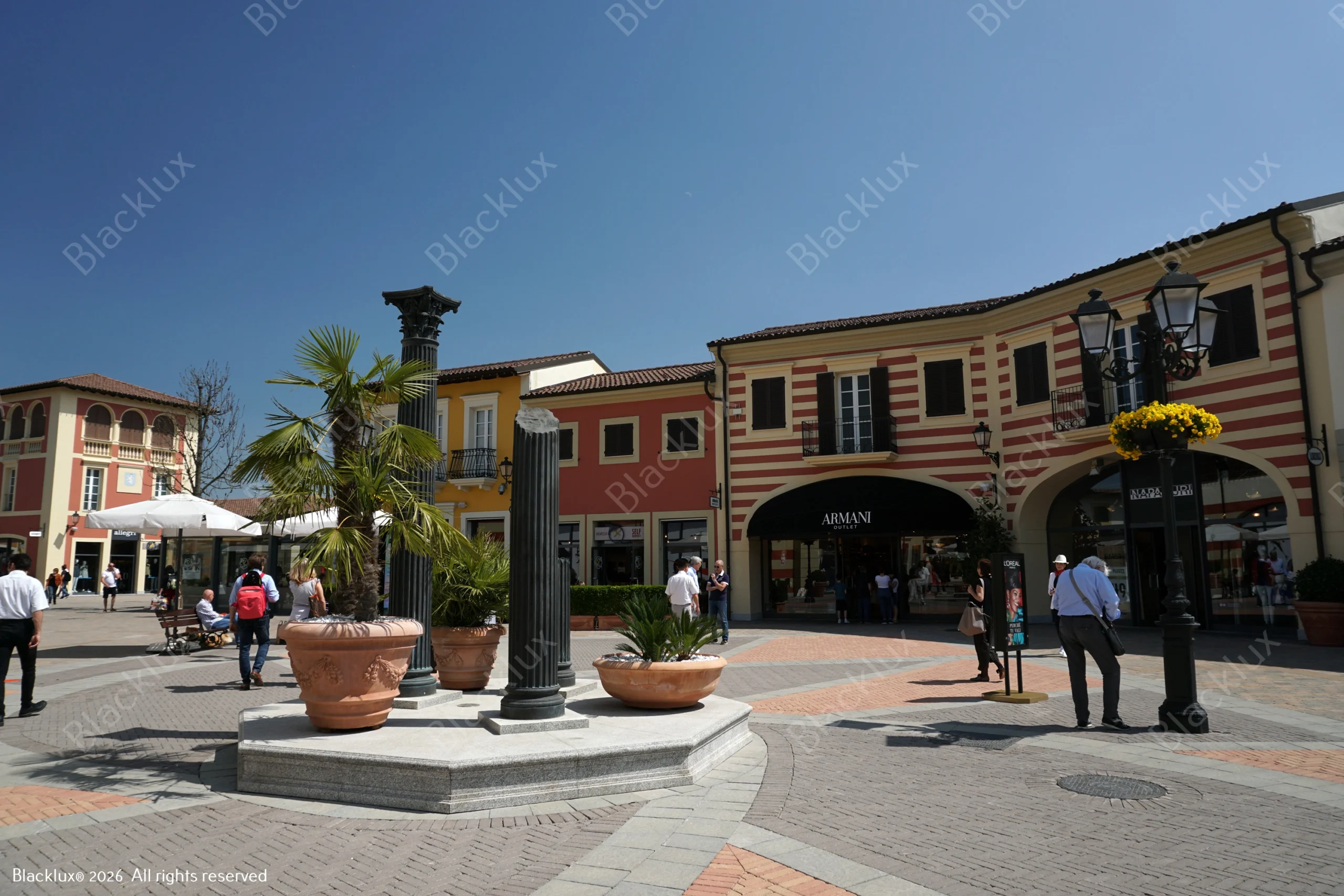 Pedestrian shopping square with terracotta buildings, arcades, potted palms and people in daylight