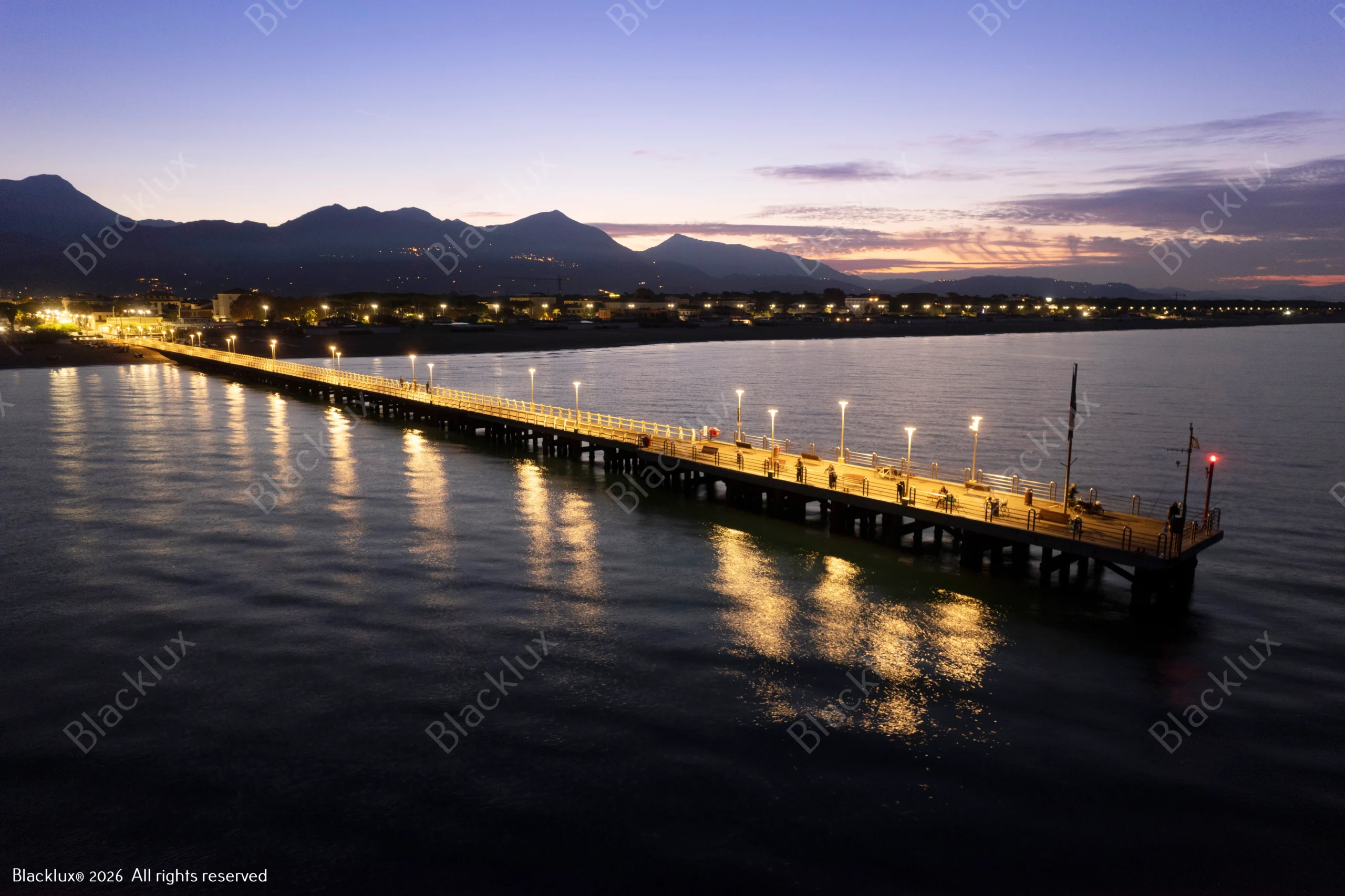 Illuminated coastal pier at dusk with mountain silhouettes and warm reflections on the sea