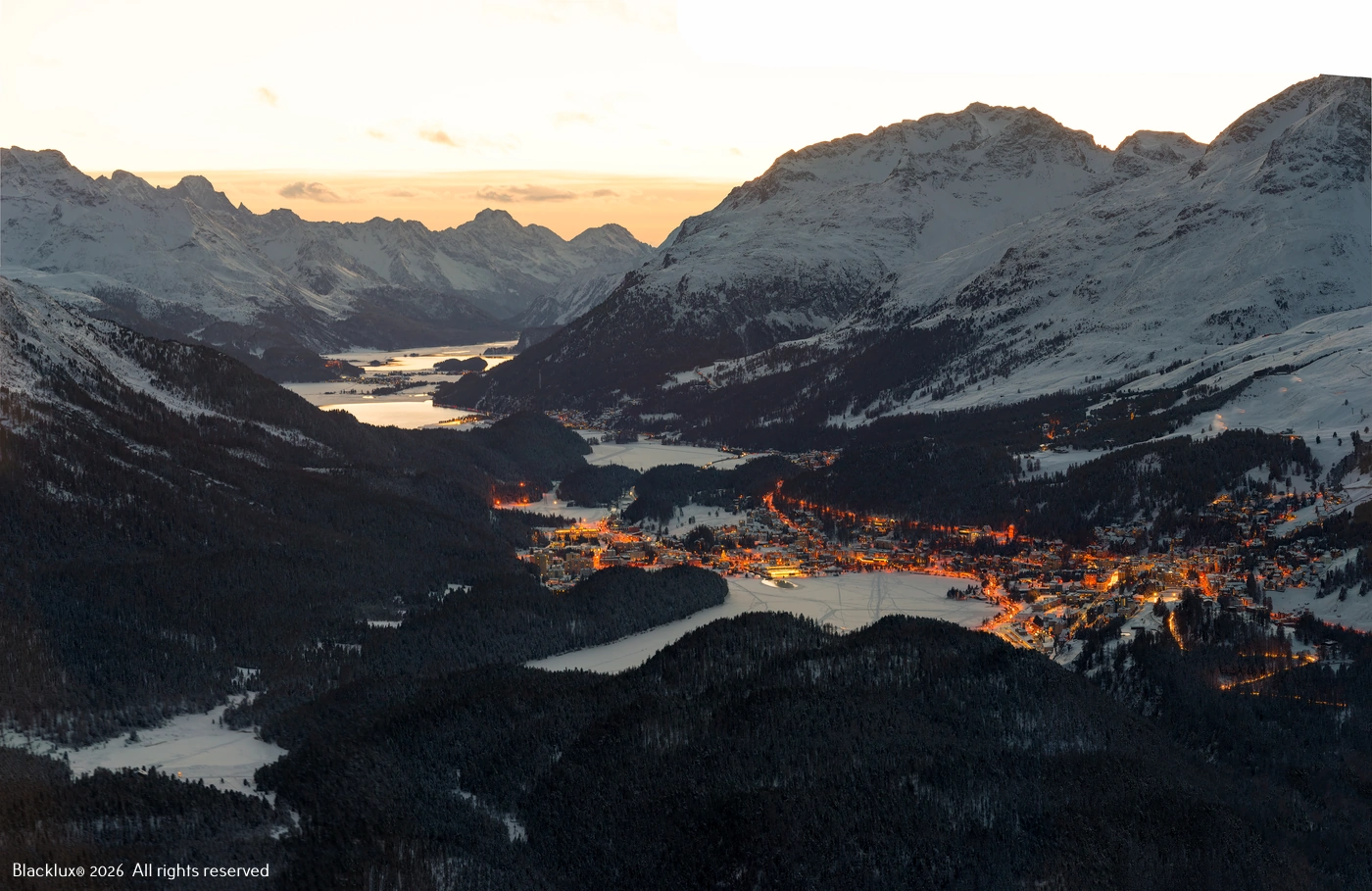 Wide alpine valley with lakes, mountain ridges and a lit settlement at dusk