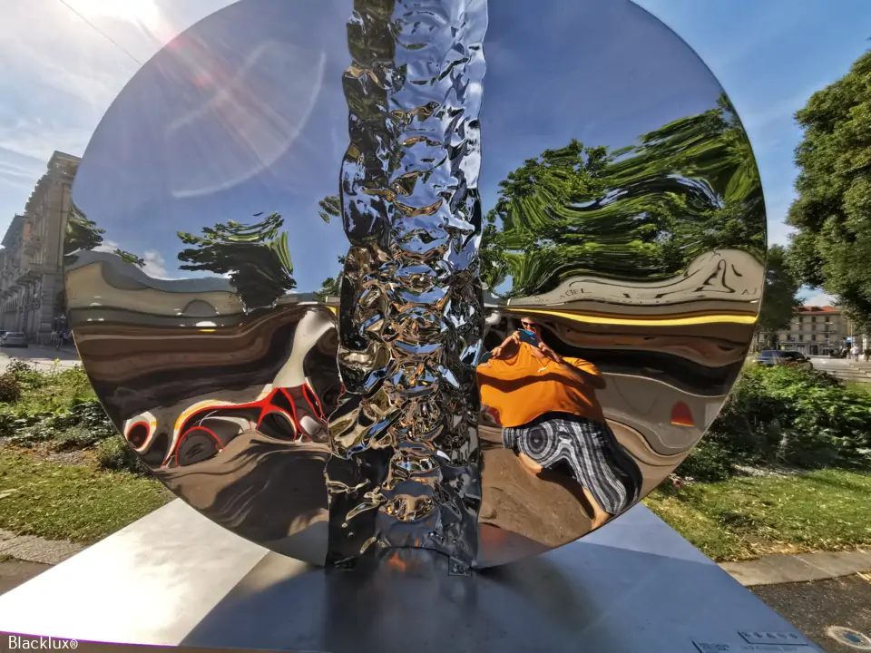 Close view of a polished circular outdoor sculpture reflecting trees, sky and surrounding buildings