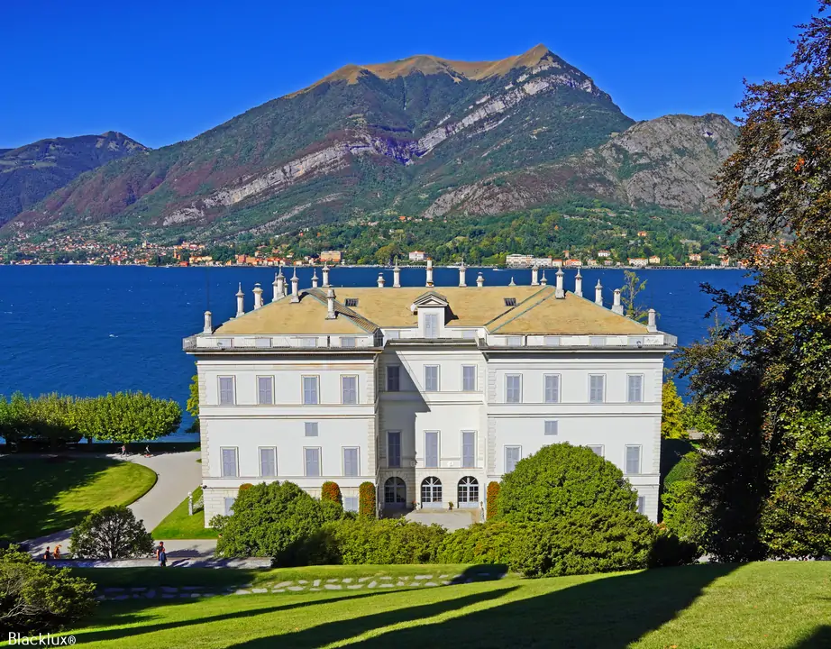 White neoclassical lakeside villa above a green lawn, with mountains across the blue lake