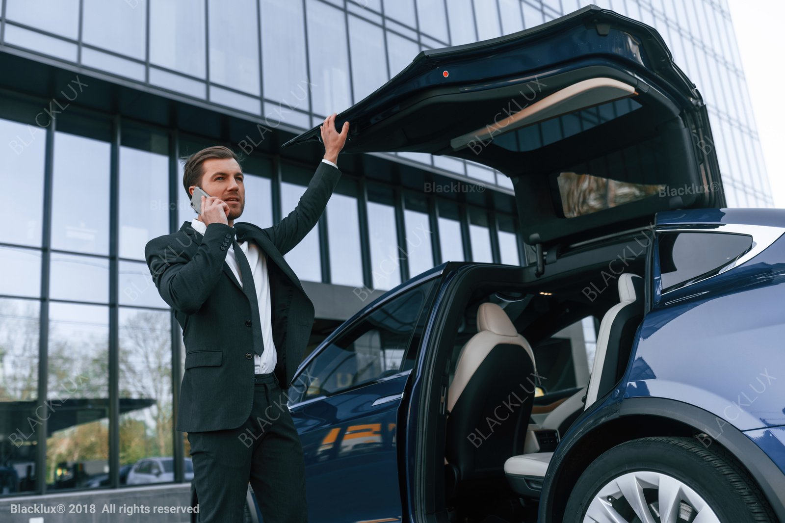 Business traveller using a phone beside a car with its boot open outside a modern building