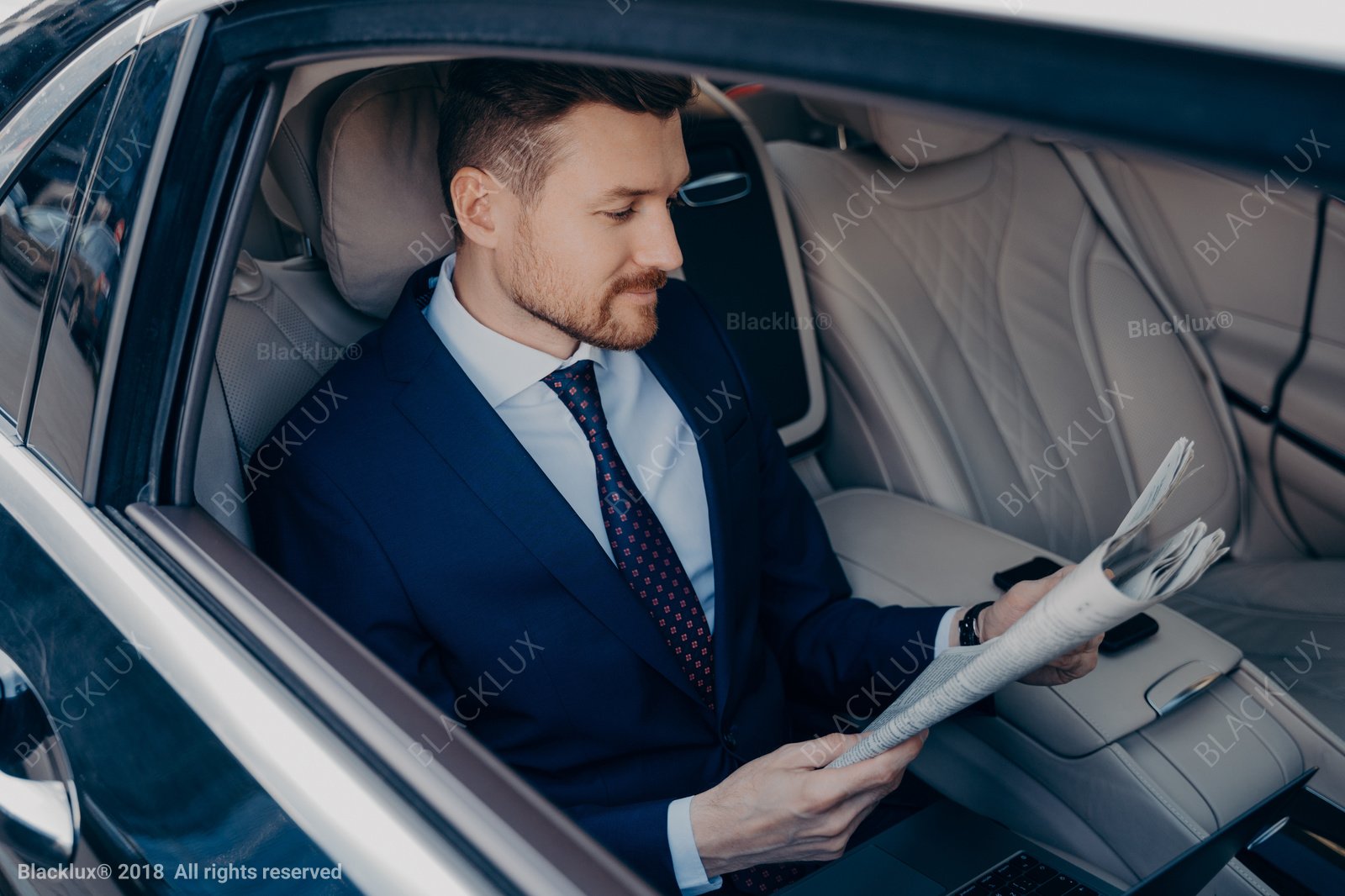 Businessman in a suit reading a newspaper in the rear seat of a car