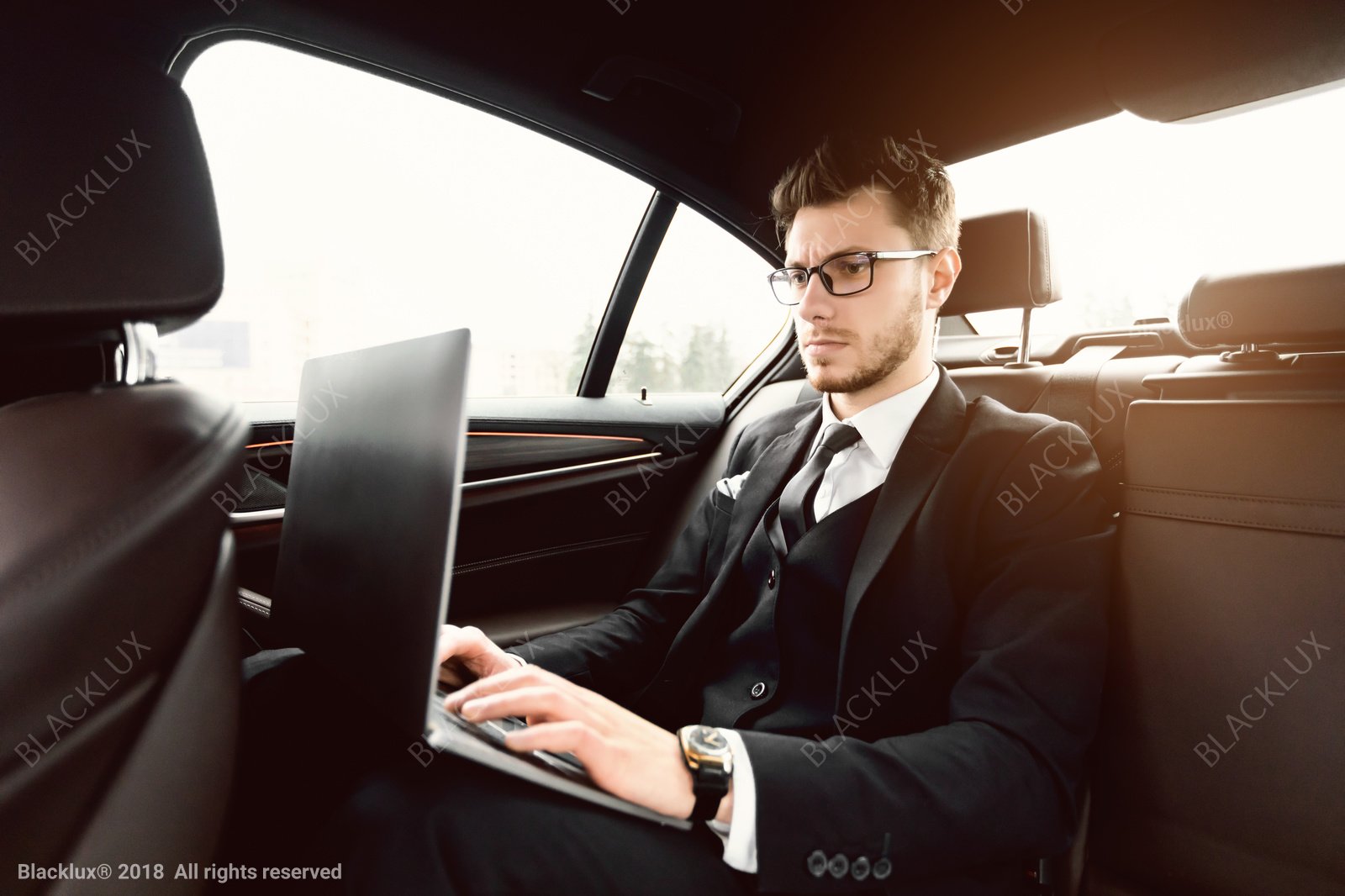 Passenger with glasses using a laptop in the rear of a dark car in daylight