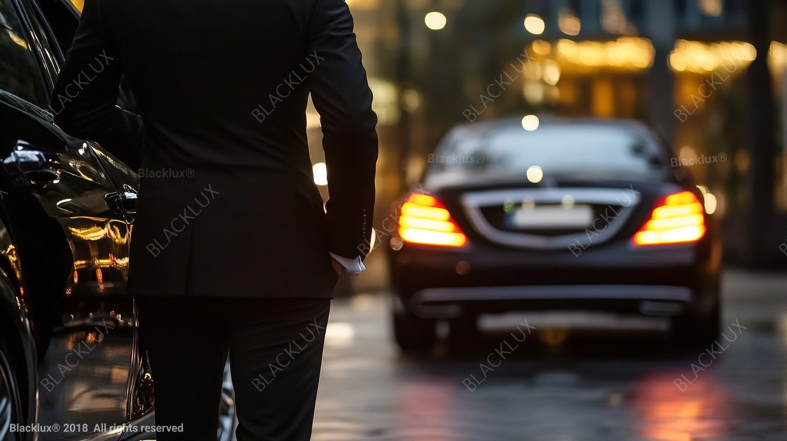 Rear view of a suited person beside a dark vehicle, with another car and warm city lights blurred ahead