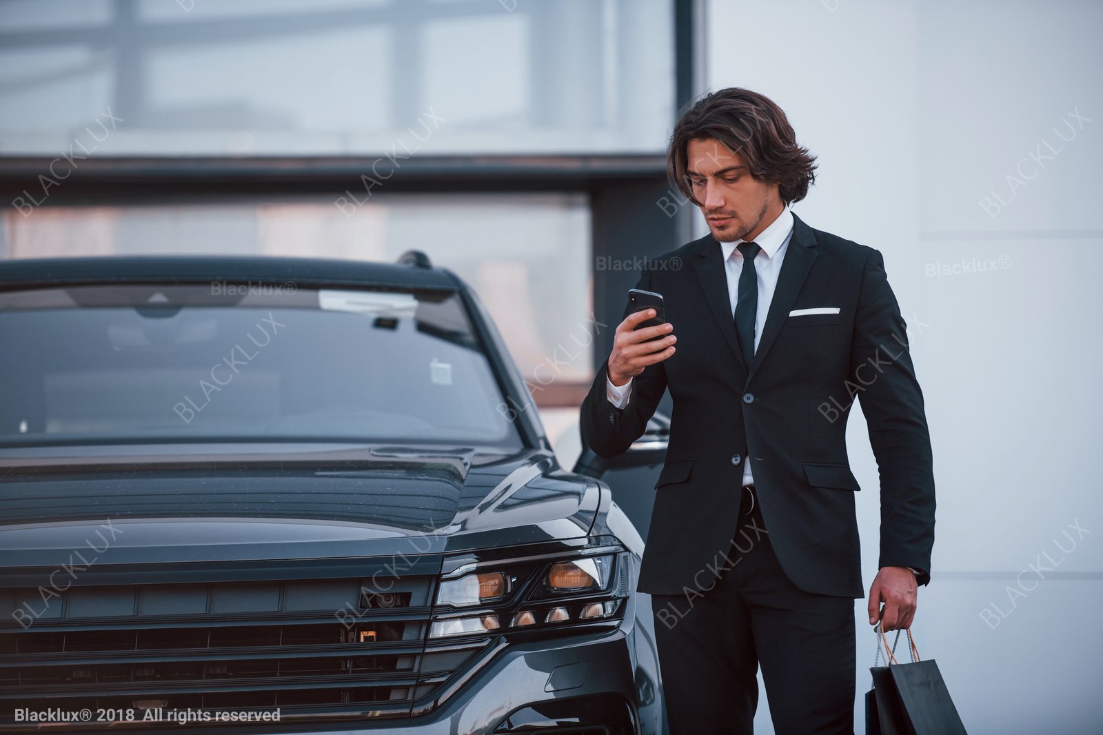 Dark-haired man in a black suit checking his phone while holding shopping bags beside a dark SUV