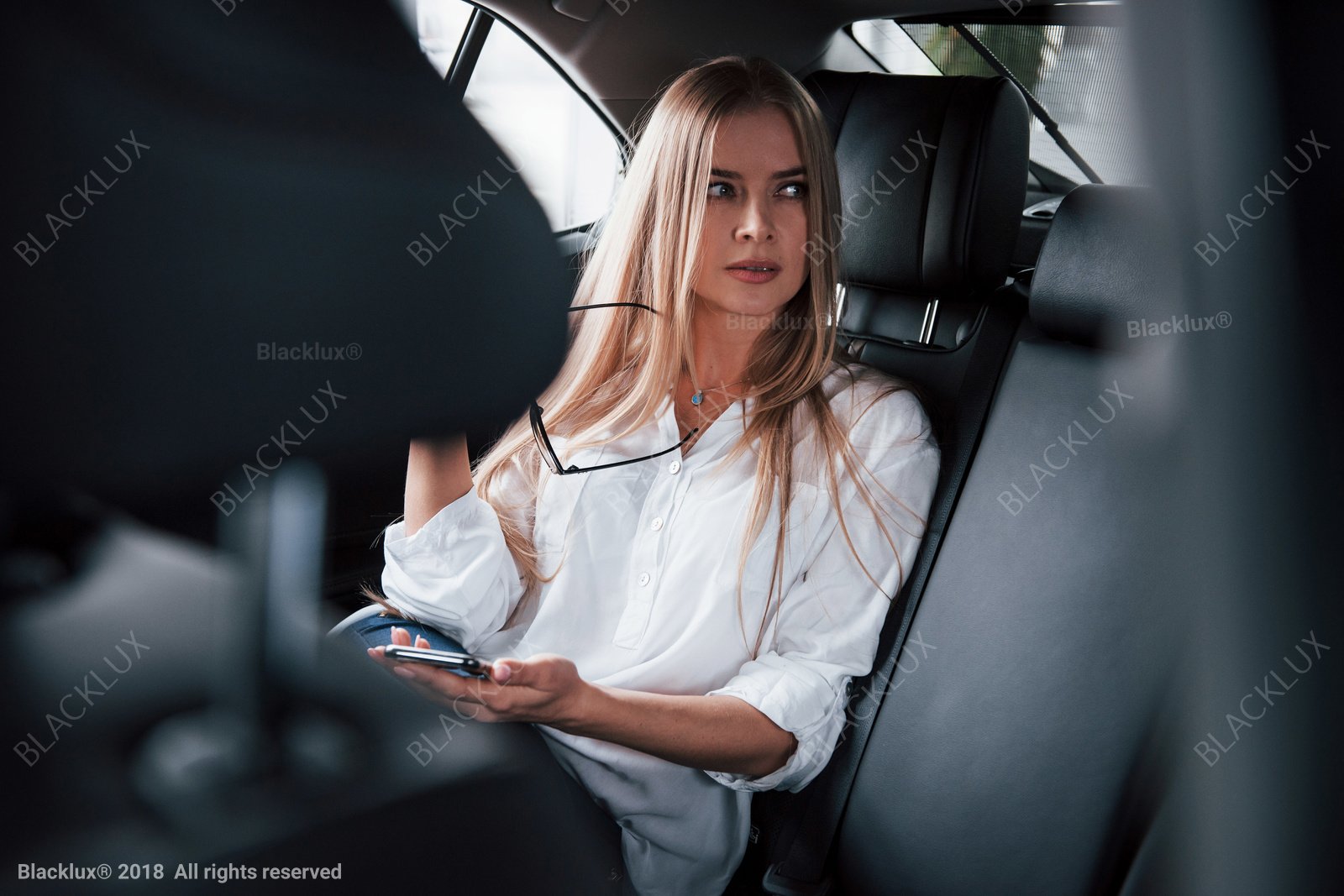 Blonde passenger in a white blouse holding a phone and glasses in a dark rear cabin
