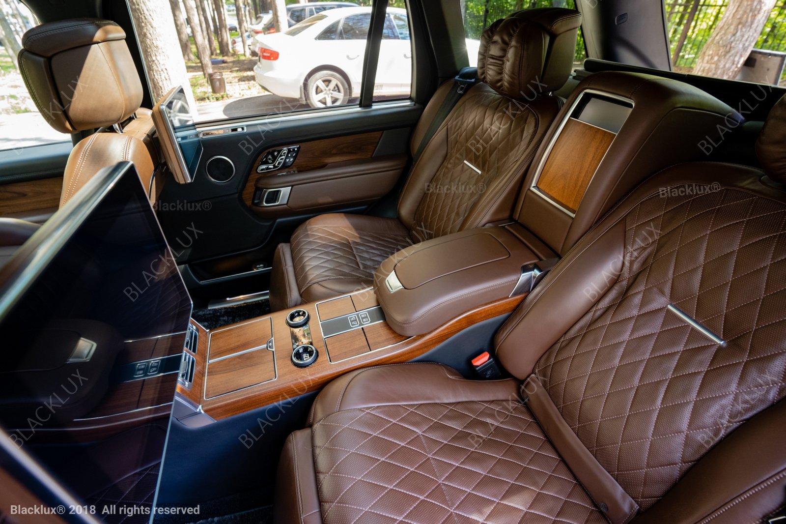 Diagonal view of brown quilted rear seats with wood trim and two headrest screens inside a vehicle