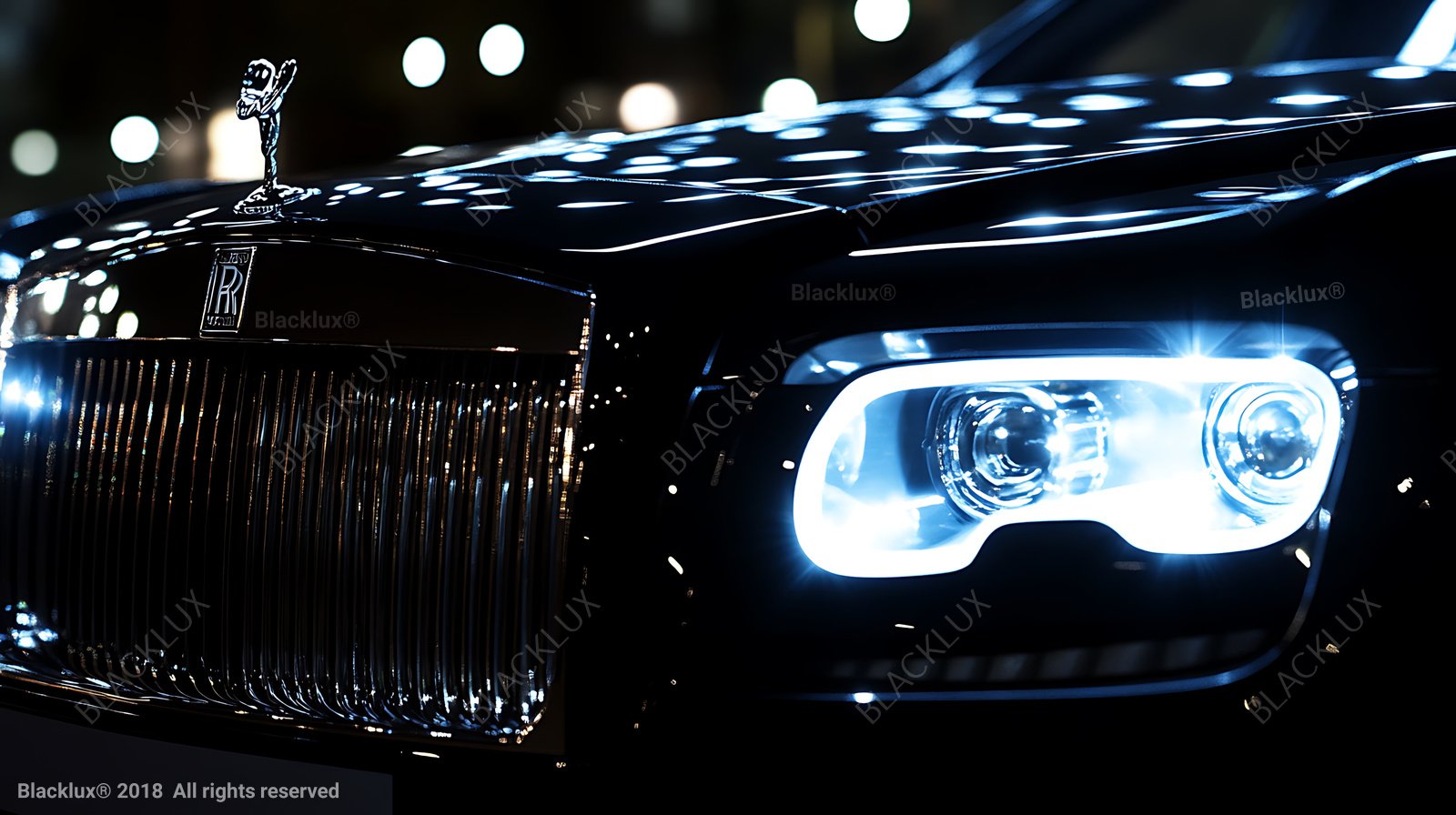 Front grille, illuminated headlight and hood ornament of a black Rolls-Royce photographed close up