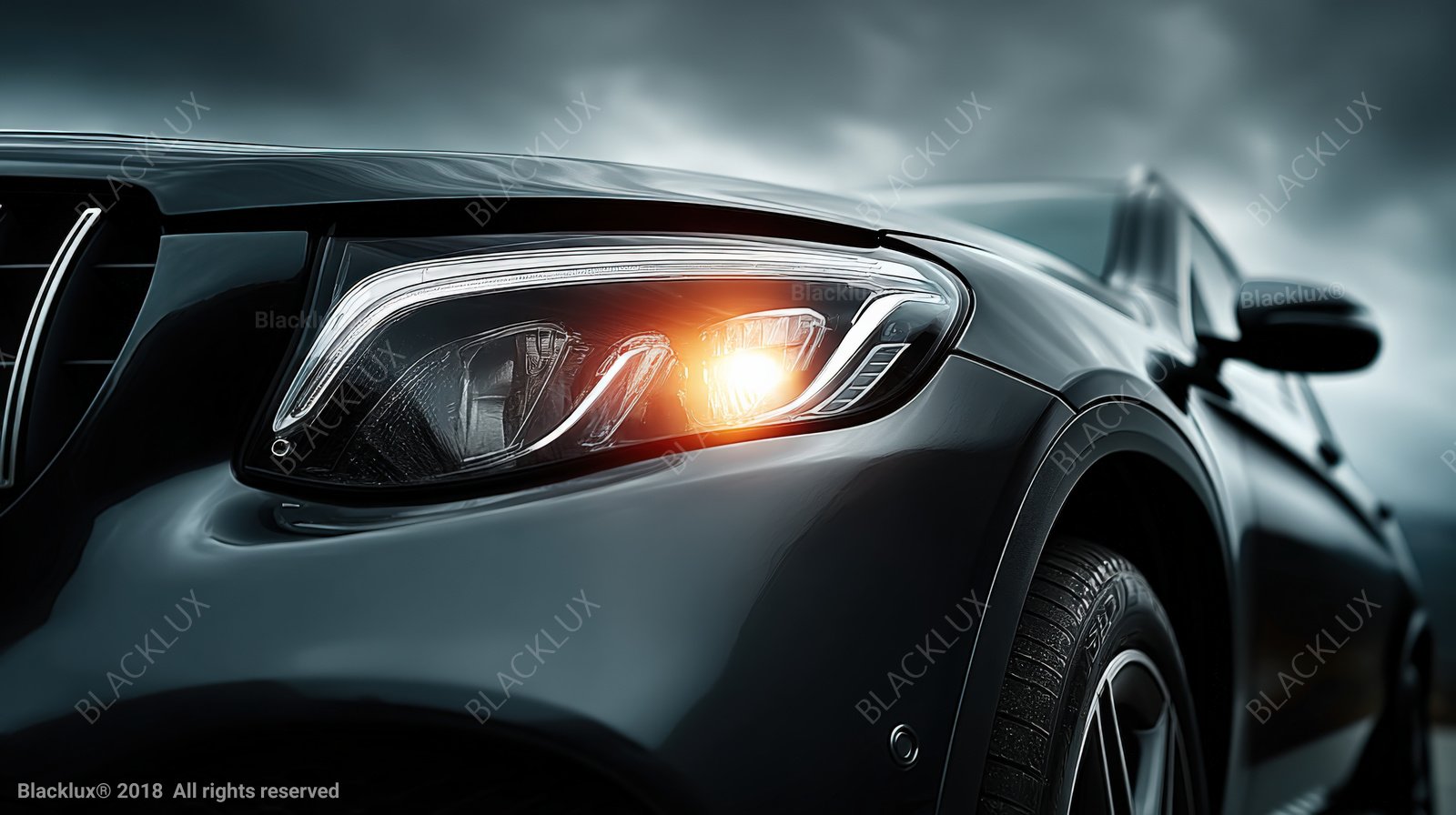 Close-up of an illuminated headlight and dark vehicle body beneath a grey-blue sky