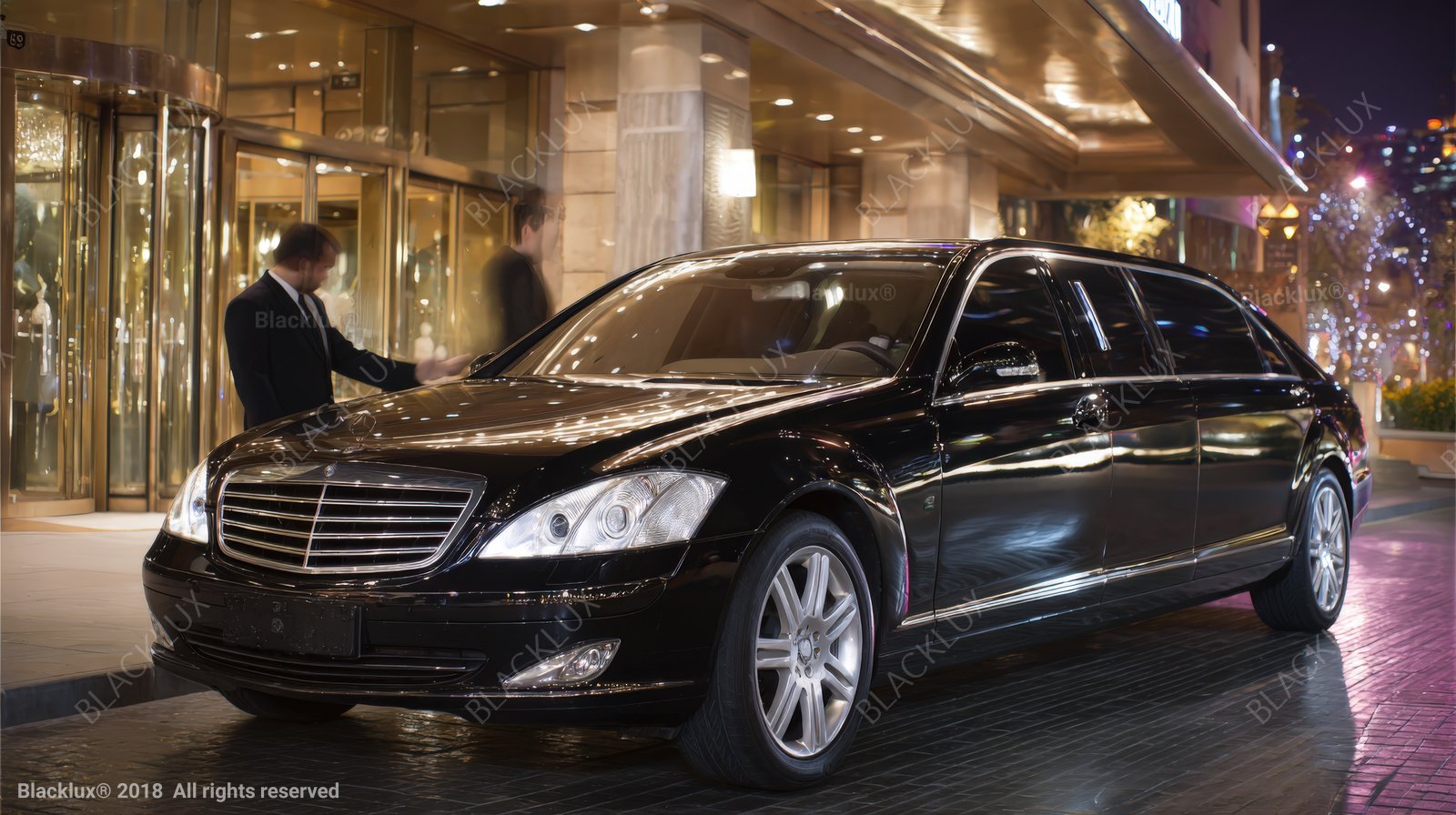 Black sedan parked beneath an illuminated hotel canopy beside a revolving entrance