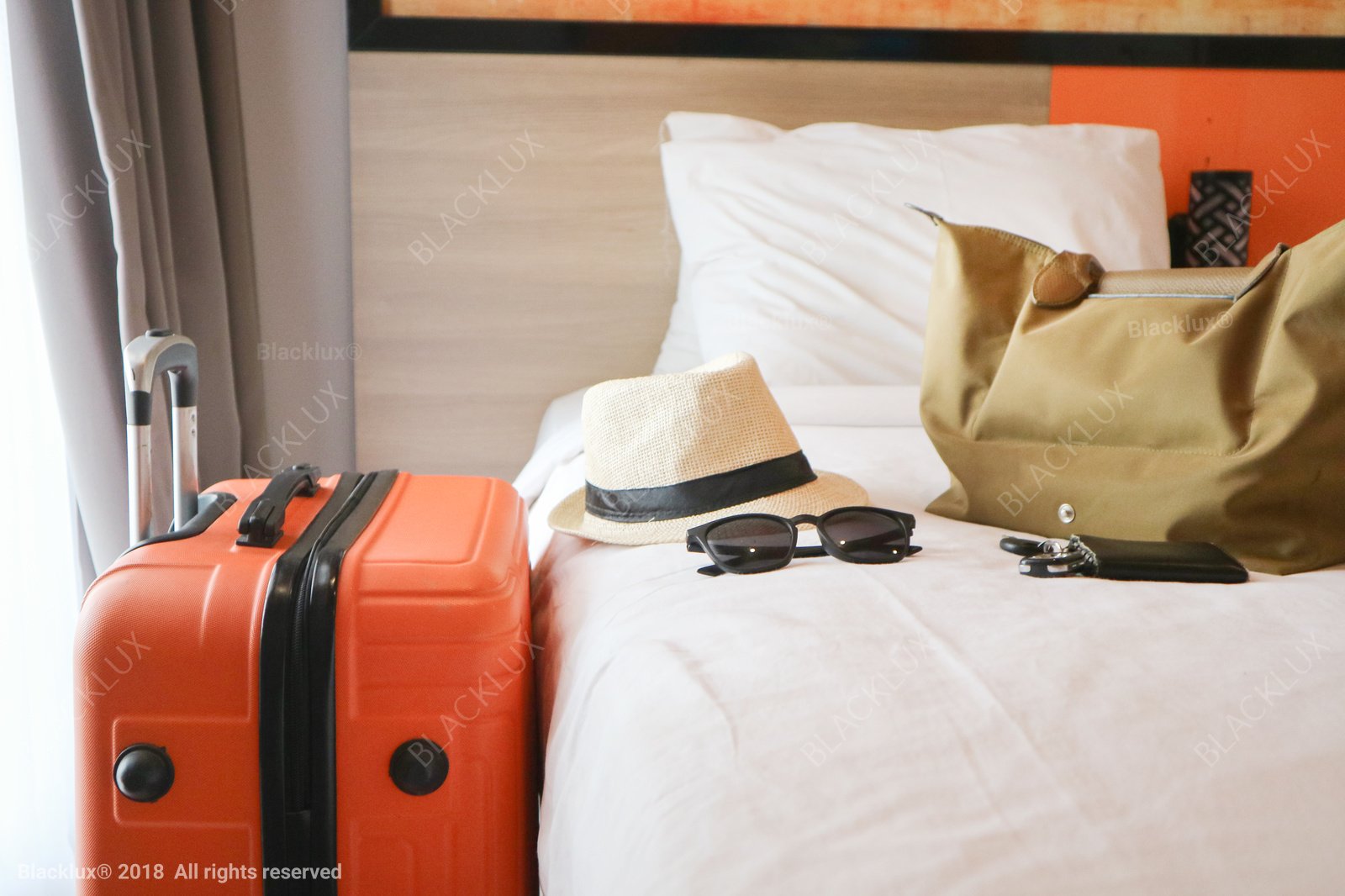 Orange suitcase beside a hotel bed with a straw hat, sunglasses and a travel bag