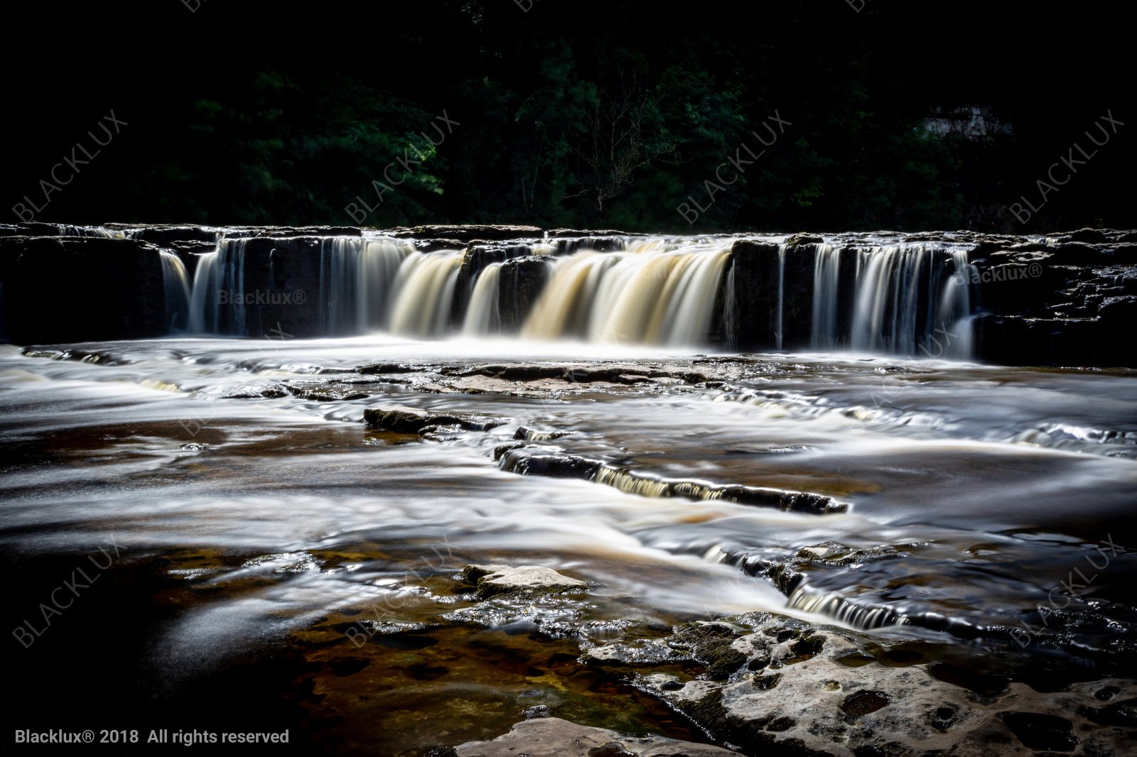 Low waterfall flowing over dark layered rocks beneath a shaded forest